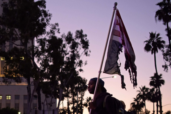 抗议示威者在洛杉矶市中心聚集,一名男子举着一面被烧过的美国国旗